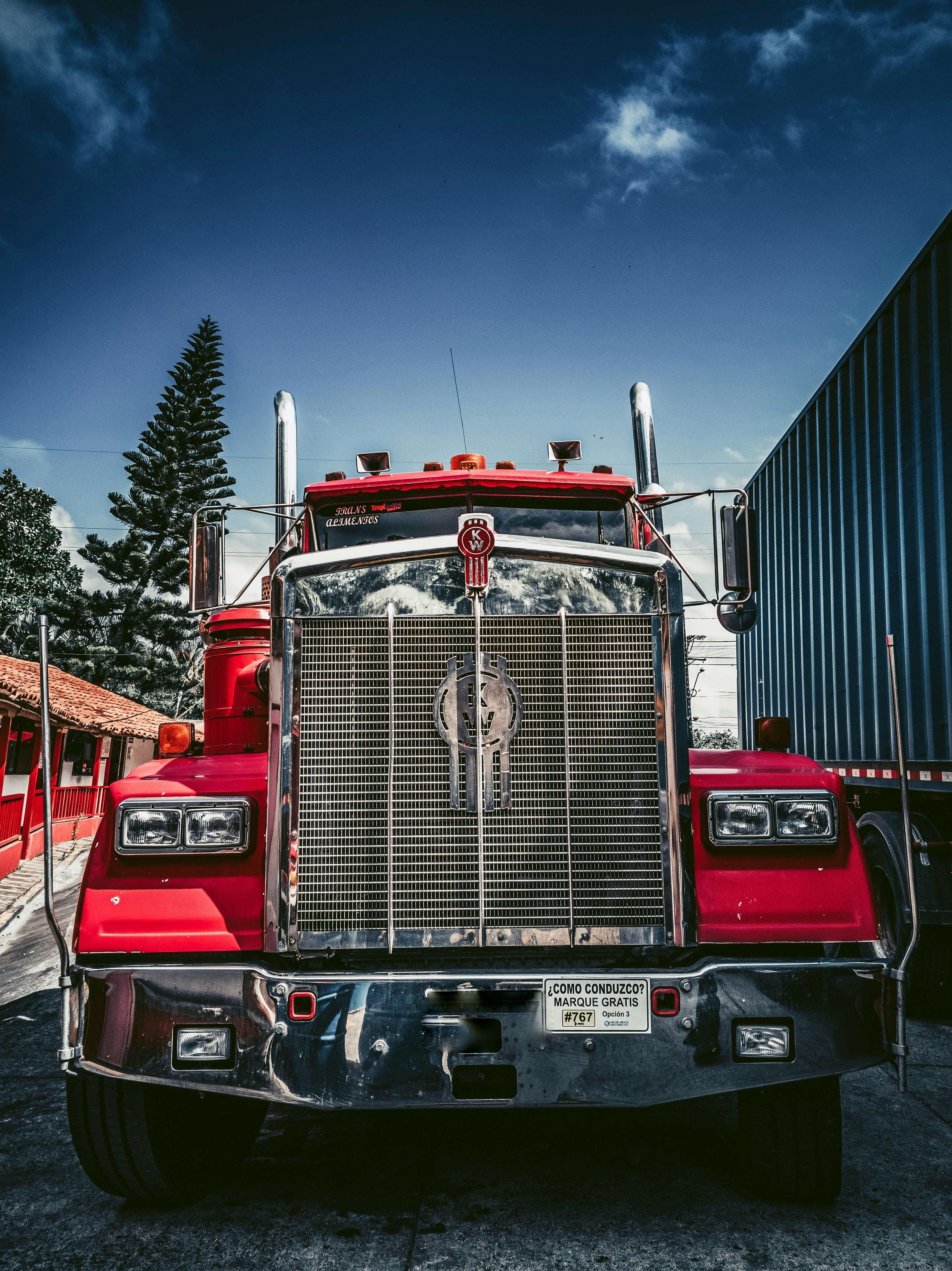 A red semi-truck, available through tow truck leasing, is parked facing the camera on a sunny day, featuring a large grille and chrome details. A blue shipping container and red building flank the truck, with pine trees and blue sky in the background.