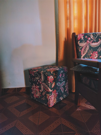 A floral patterned cube-shaped ottoman is placed on a tiled floor beside a matching upholstered chair. The scene is softly lit by natural light filtering through golden-yellow curtains, casting gentle shadows on a neutral-colored wall.