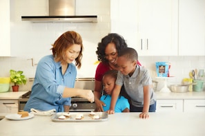 A family of four in a kitchen preparing food together. The adults and children are focused on baking, with a tray of ingredients on the counter. The kitchen is modern and clean, with white cabinets and various utensils and appliances visible in the background.