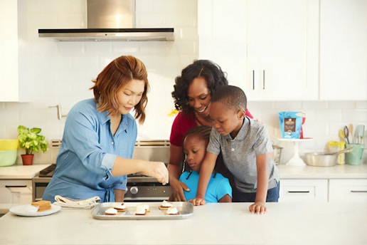 A family of four in a kitchen preparing food together. The adults and children are focused on baking, with a tray of ingredients on the counter. The kitchen is modern and clean, with white cabinets and various utensils and appliances visible in the background.
