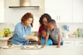A family of four in a kitchen preparing food together. The adults and children are focused on baking, with a tray of ingredients on the counter. The kitchen is modern and clean, with white cabinets and various utensils and appliances visible in the background.