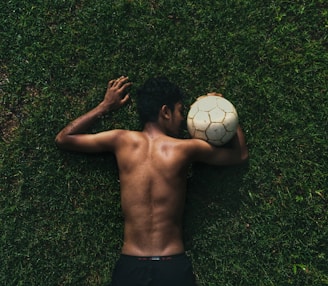 A person lies face down on a grassy field. The individual is shirtless and holds a worn-out soccer ball close to their head.