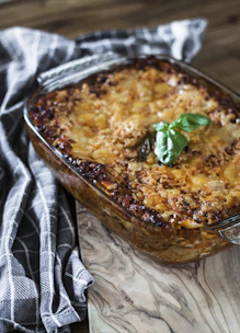 A close-up of a freshly baked lasagna in a rectangular glass dish. The top is golden brown with visible layers of cheese and tomato sauce, garnished with a sprig of fresh basil. It rests on a marbled countertop beside a folded checkered kitchen towel.