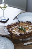 A freshly baked lasagna in a glass dish rests on a wooden board, garnished with a sprig of basil and sprinkled with slices of parmesan cheese. Beside it, there is an empty plate and a knife, suggesting preparation for serving. A glass of white wine sits on a coaster, adding an elegant touch to the scene.