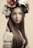 A young woman with long brown hair wears a traditional embroidered blouse and a floral wreath on her head, featuring blossoms in colors like red, white, and green. The overall appearance is traditional and serene.