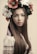 A young woman with long brown hair wears a traditional embroidered blouse and a floral wreath on her head, featuring blossoms in colors like red, white, and green. The overall appearance is traditional and serene.