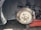 A close-up view of a car's wheel area, showing a removed tire and a visible brake disc with a rusty caliper. Part of a red hydraulic jack is seen supporting the car. The suspension system, including the spring and shock absorber, is visible.
