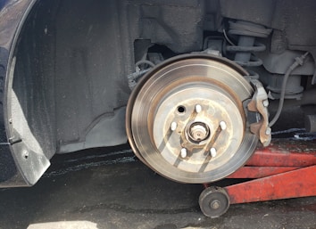 A close-up view of a car's wheel area, showing a removed tire and a visible brake disc with a rusty caliper. Part of a red hydraulic jack is seen supporting the car. The suspension system, including the spring and shock absorber, is visible.