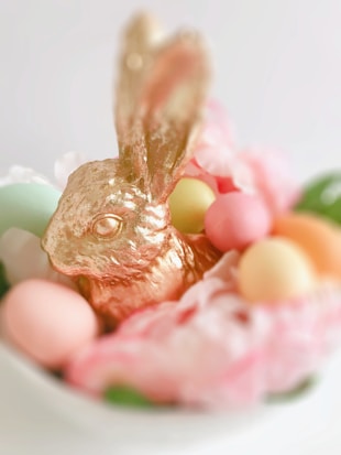 A golden, textured rabbit figurine surrounded by pastel-colored Easter eggs and delicate pink flowers. Soft lighting enhances the pastel hues, creating a gentle and whimsical atmosphere.
