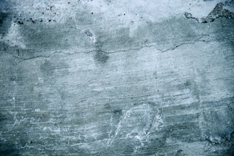 A textured concrete wall with visible cracks and discoloration. The surface shows signs of wear, including uneven patches and faint lines across the greyish surface.