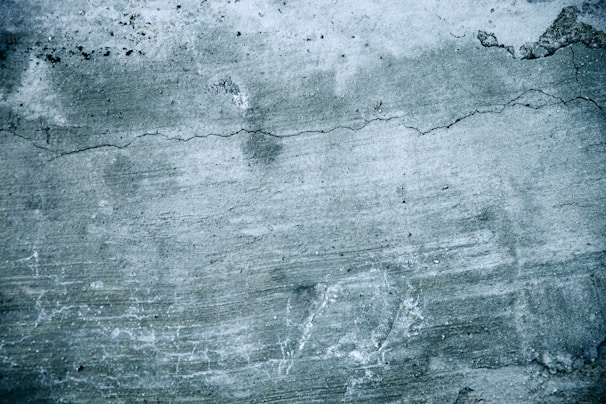 A textured concrete wall with visible cracks and discoloration. The surface shows signs of wear, including uneven patches and faint lines across the greyish surface.