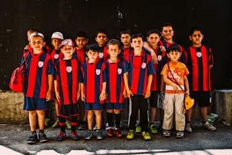 A group of young boys dressed in matching red and black striped sports jerseys pose together. Most of them are wearing sports shorts and athletic shoes, while one child stands out in a colorful T-shirt and cargo pants. The boys display a range of emotions from serious to relaxed expressions, standing against a dark background.