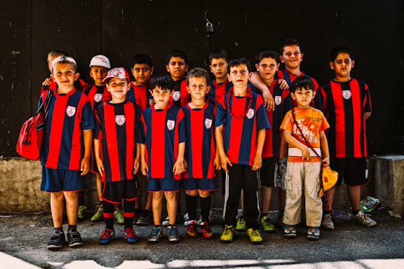A group of young boys dressed in matching red and black striped sports jerseys pose together. Most of them are wearing sports shorts and athletic shoes, while one child stands out in a colorful T-shirt and cargo pants. The boys display a range of emotions from serious to relaxed expressions, standing against a dark background.