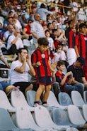 A young boy in a red and black striped sports outfit stands on a stadium seat, appearing excited. Around him, a crowd of seated spectators, including adults and children, show various expressions of enthusiasm and anticipation. The seating area is filled, suggesting a lively event atmosphere.