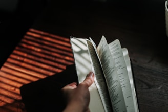 A hand flipping through the pages of an open book, with shadows and light filtering through blinds onto a wooden surface, creating a warm and contemplative atmosphere.