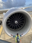 A large airplane engine dominates the frame while a person wearing a high-visibility jacket stands with arms outstretched underneath. The sky is partly cloudy in the background, and the airplane extends into the distance on one side.
