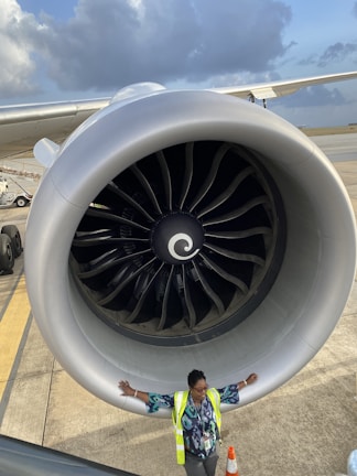 A large airplane engine dominates the frame while a person wearing a high-visibility jacket stands with arms outstretched underneath. The sky is partly cloudy in the background, and the airplane extends into the distance on one side.