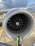 A large airplane engine dominates the frame while a person wearing a high-visibility jacket stands with arms outstretched underneath. The sky is partly cloudy in the background, and the airplane extends into the distance on one side.