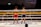 Two Muay Thai fighters stand in a boxing ring facing each other, wearing traditional headbands and gloves. The fighter on the left wears red shorts, while the one on the right wears blue. The ropes of the ring are adorned with signage, and the background is filled with spectators observing the match.