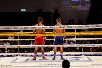 Two Muay Thai fighters stand in a boxing ring facing each other, wearing traditional headbands and gloves. The fighter on the left wears red shorts, while the one on the right wears blue. The ropes of the ring are adorned with signage, and the background is filled with spectators observing the match.