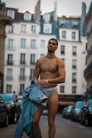 A shirtless person stands confidently in a street lined with tall buildings, holding denim clothing. The image is set in an urban environment, with cars parked along the sides and classic European architecture in the background.