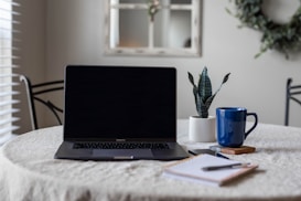 A laptop with a dark screen is placed on a round table covered with a white tablecloth. To the right of the laptop is a small potted plant with dark green leaves and a blue coffee mug on a coaster. A notebook with a pen rests in front of the laptop. In the background, the room features a blurred reflection of a window in a mirror and a decorative wreath on the wall.
