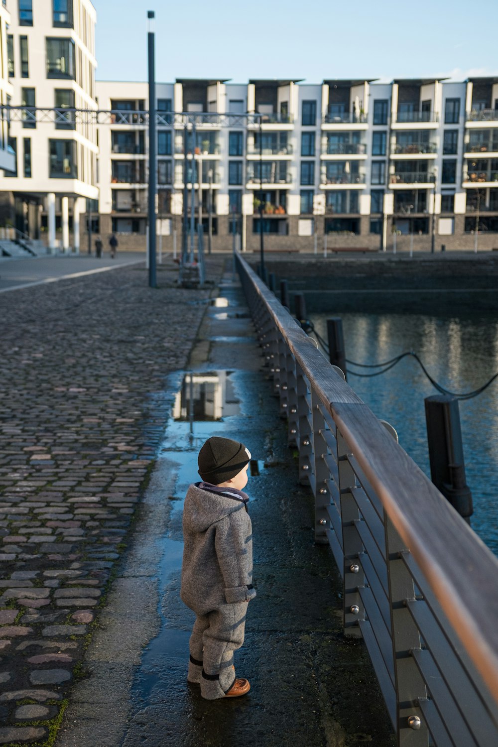 person in brown coat standing on gray concrete pavement near body of water during daytime