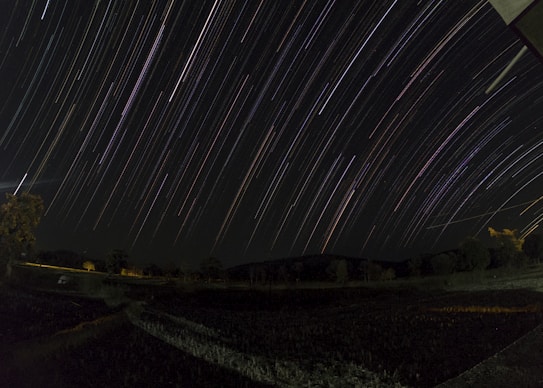 A long-exposure photograph captures star trails across a dark night sky, with trees and distant hills visible in the landscape. The paths of the stars create curved lines of varying colors and brightness, suggesting a sense of movement and time passing.