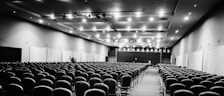 A large, empty conference hall with numerous rows of chairs neatly arranged, facing a stage at the far end. The ceiling is fitted with lights, and curtains line the side walls.