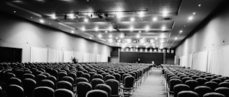 A large, empty conference hall with numerous rows of chairs neatly arranged, facing a stage at the far end. The ceiling is fitted with lights, and curtains line the side walls.