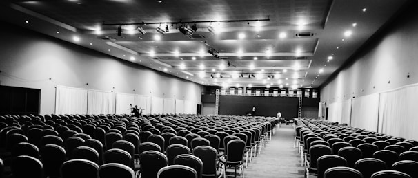 A large, empty conference hall with numerous rows of chairs neatly arranged, facing a stage at the far end. The ceiling is fitted with lights, and curtains line the side walls.
