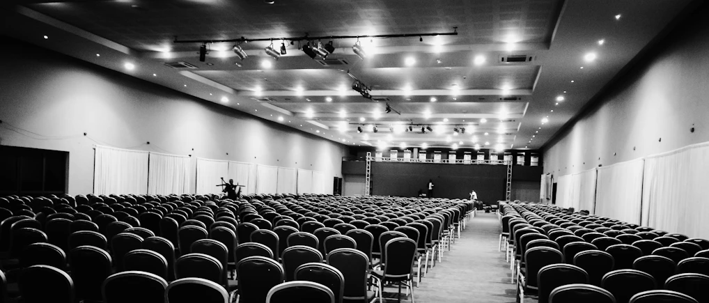 A bright, spacious conference hall filled with rows of chairs and a large stage at the front.