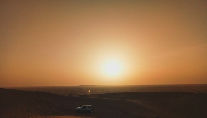 A sunset over a vast desert landscape with a vehicle parked on the sand. The sun casts a warm, golden light across the sky, with the horizon visible in the distance.