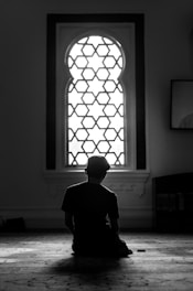 A person seated on the floor with their back to the camera, gazing towards an intricately designed window that lets in bright light. The setting suggests a serene and contemplative mood, possibly inside a place of worship.