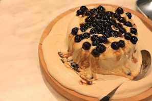 A round dessert covered with black tapioca pearls is placed on a wooden plate. The dessert appears creamy and is partially melted, with a silver spoon resting on the side.