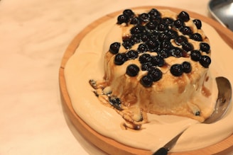 A round dessert covered with black tapioca pearls is placed on a wooden plate. The dessert appears creamy and is partially melted, with a silver spoon resting on the side.