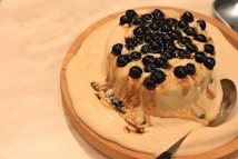 A round dessert covered with black tapioca pearls is placed on a wooden plate. The dessert appears creamy and is partially melted, with a silver spoon resting on the side.