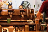 An industrial-style coffee machine with multiple gauges and levers is featured with a barista preparing a cup of coffee. Various coffee-related items like cups, pitchers, and a cloth are scattered around. The background shows blurred images of mannequins and plants.