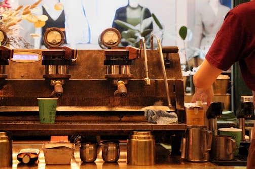 An industrial-style coffee machine with multiple gauges and levers is featured with a barista preparing a cup of coffee. Various coffee-related items like cups, pitchers, and a cloth are scattered around. The background shows blurred images of mannequins and plants.