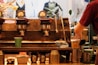 An industrial-style coffee machine with multiple gauges and levers is featured with a barista preparing a cup of coffee. Various coffee-related items like cups, pitchers, and a cloth are scattered around. The background shows blurred images of mannequins and plants.