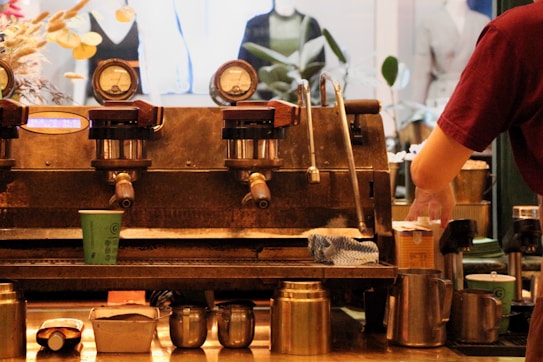 An industrial-style coffee machine with multiple gauges and levers is featured with a barista preparing a cup of coffee. Various coffee-related items like cups, pitchers, and a cloth are scattered around. The background shows blurred images of mannequins and plants.