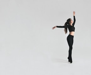 A dancer in a black outfit poses gracefully against a minimalist light grey background. The dancer is mid-motion, arms elegantly extended, showcasing a dynamic yet poised posture.