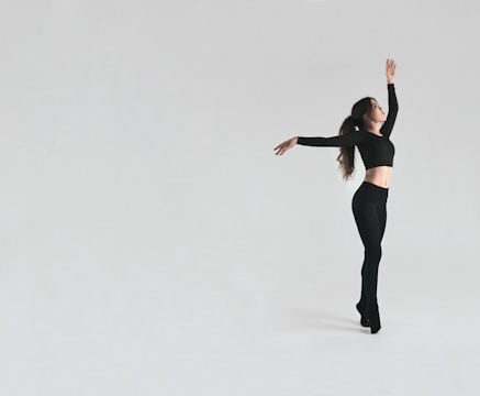 A dancer in a black outfit poses gracefully against a minimalist light grey background. The dancer is mid-motion, arms elegantly extended, showcasing a dynamic yet poised posture.