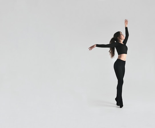 A dancer in a black outfit poses gracefully against a minimalist light grey background. The dancer is mid-motion, arms elegantly extended, showcasing a dynamic yet poised posture.