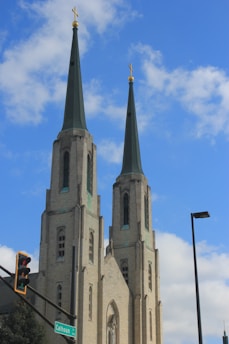 Two tall church towers with green-tipped spires and gold crosses rise against a backdrop of a clear blue sky with a few clouds. A traffic light and street sign for Calhoun St are visible at the corner of the image, along with a street lamp.