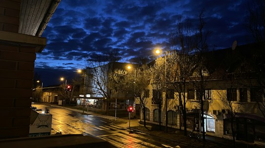 A quiet, dimly lit street scene at dusk, with buildings lining the road and streetlights casting a soft glow. The sky is a deep blue, dotted with dark clouds. Bare trees stand on the sidewalk, and reflections from the wet road surface are visible. Traffic lights are seen in the distance, and the image conveys a sense of calm and tranquility.