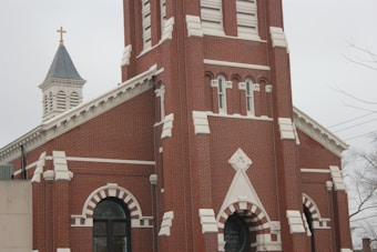 A red brick church with white architectural detailing features a tall bell tower and a pointed steeple with a golden cross on top. The building has arched windows and a decorative entrance, emphasizing its traditional design.