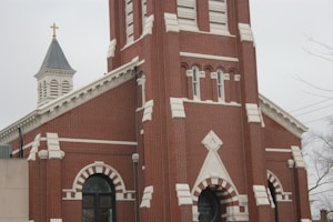 A red brick church with white architectural detailing features a tall bell tower and a pointed steeple with a golden cross on top. The building has arched windows and a decorative entrance, emphasizing its traditional design.