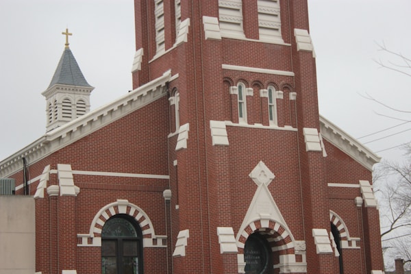 A red brick church with white architectural detailing features a tall bell tower and a pointed steeple with a golden cross on top. The building has arched windows and a decorative entrance, emphasizing its traditional design.