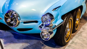 A vintage-style blue car with a custom design featuring three wheels on one side. The car body is a shiny blue with chrome accents, including the distinctive front bumper and headlights. The wheels have golden rims, adding a touch of elegance to the vehicle.